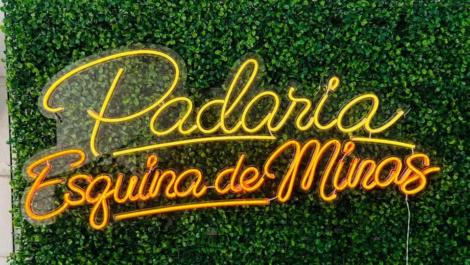 Painel neon LED em script "Padaria Esquina de Minas" aceso sobre parede verde